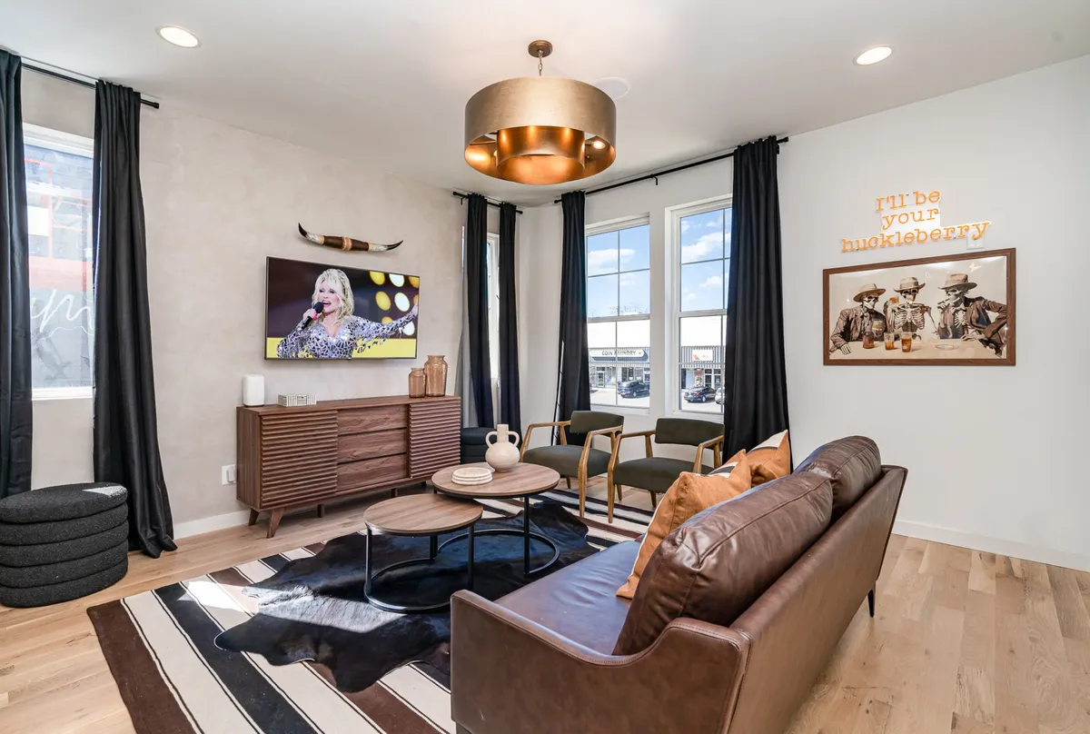 A stylish lounge area with a comfortable sofa and seating arrangement, a coffee table with decorative items, and a television displaying a live performance. Artwork adorns the wall, enhancing the room’s creative and lively ambiance.