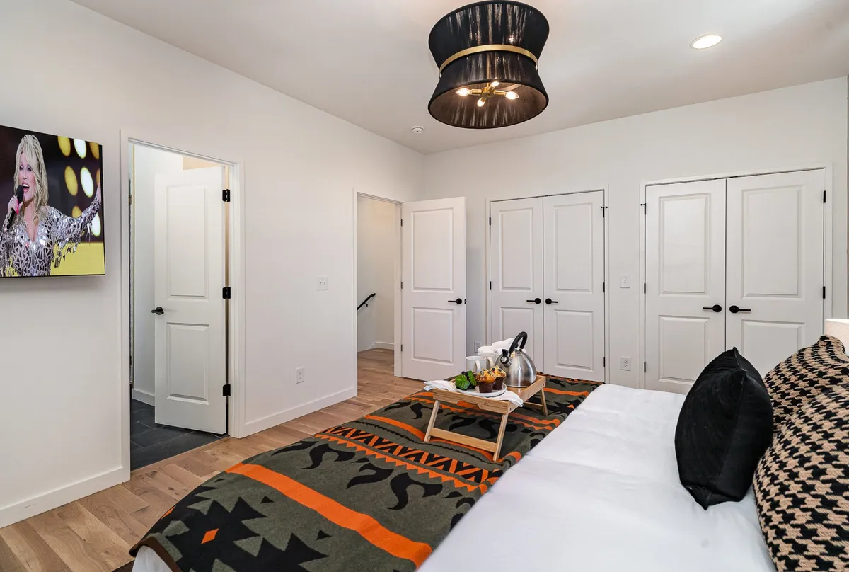 A spacious, stylish bedroom with a large bed adorned with a decorative throw pillow, a geometric patterned rug, a wall-mounted television, and a striking modern pendant light. The room is bathed in natural light, creating a cozy yet upscale ambiance.