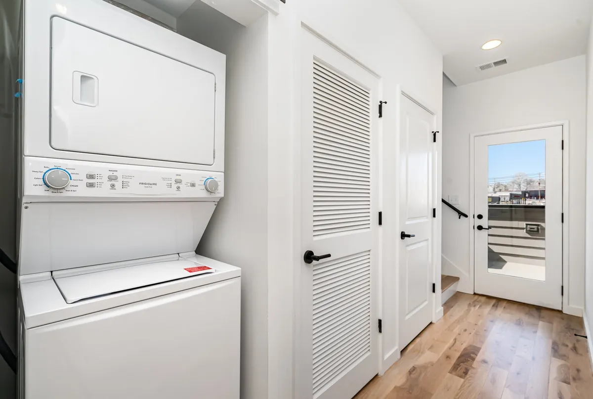 A tidy interior utility space with a stacked washer and dryer, light walls, wooden flooring, and a door leading directly outside, making it easy to manage household tasks and access the exterior.