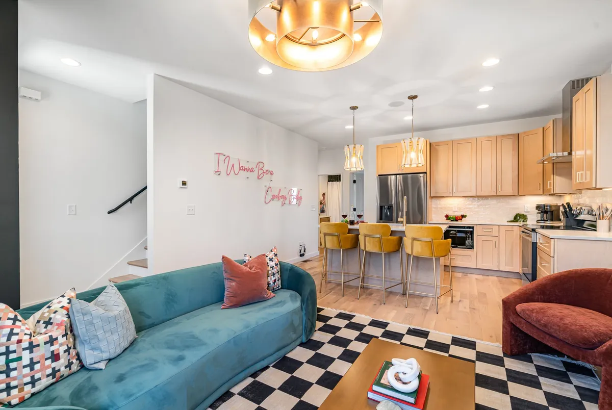 A vibrant open-concept living and kitchen space featuring a turquoise sofa, checkered rug, golden bar stools, and a stylish kitchen area with modern appliances. Decorative wall text adds a playful, creative flair, while the layout naturally encourages social gatherings and interaction.