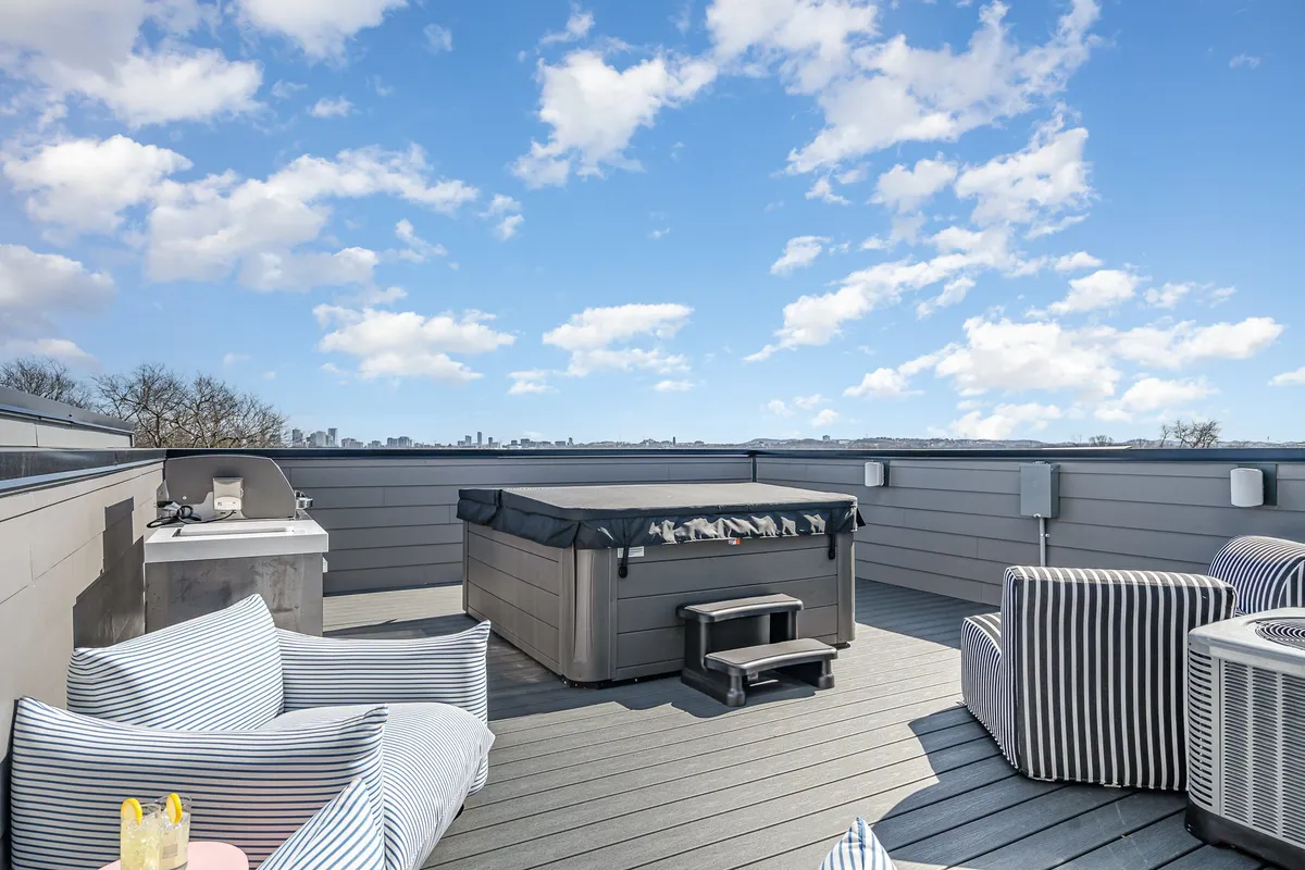 A cozy outdoor setting on a rooftop deck with a hot tub, striped chairs, and an outdoor grill. The scene transitions beautifully from sunset to nighttime, emphasizing both relaxation and entertainment.