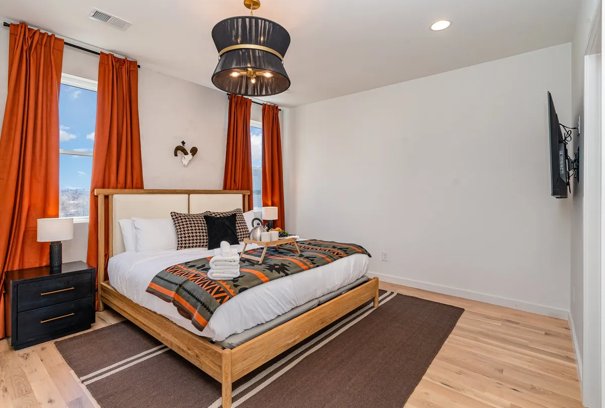 A contemporary bedroom featuring a bed with a patterned blanket, bold orange curtains, a nightstand with a lamp, and a TV mounted on the wall. The space is bright and inviting, with modern accents that make it suitable for a guest room setting.
