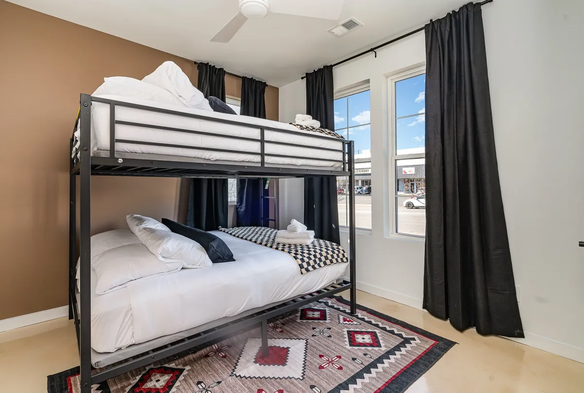 A playful bunk room with a sturdy bunk bed, a soft textured rug, and large windows that fill the room with natural light. The space is designed to be comfortable and fun, ideal for children or groups sharing a room.